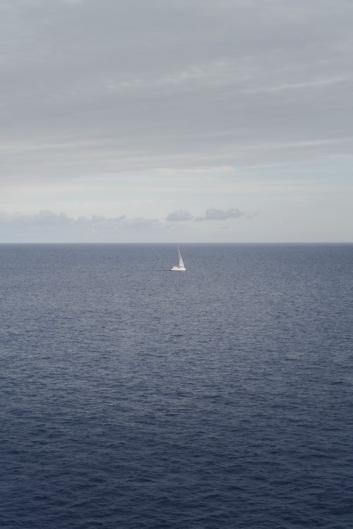 Free A lone sailboat navigates calm waters of the open ocean with an overcast sky overhead. Stock Photo