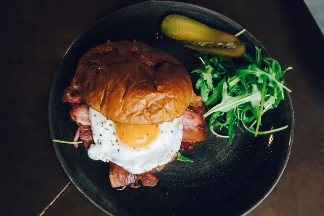 Fried egg and smoked bacon in a toasted brioche roll, plated with rocket and a dill pickle. Photo by Medine Dilek Kizmaz on Pexels.