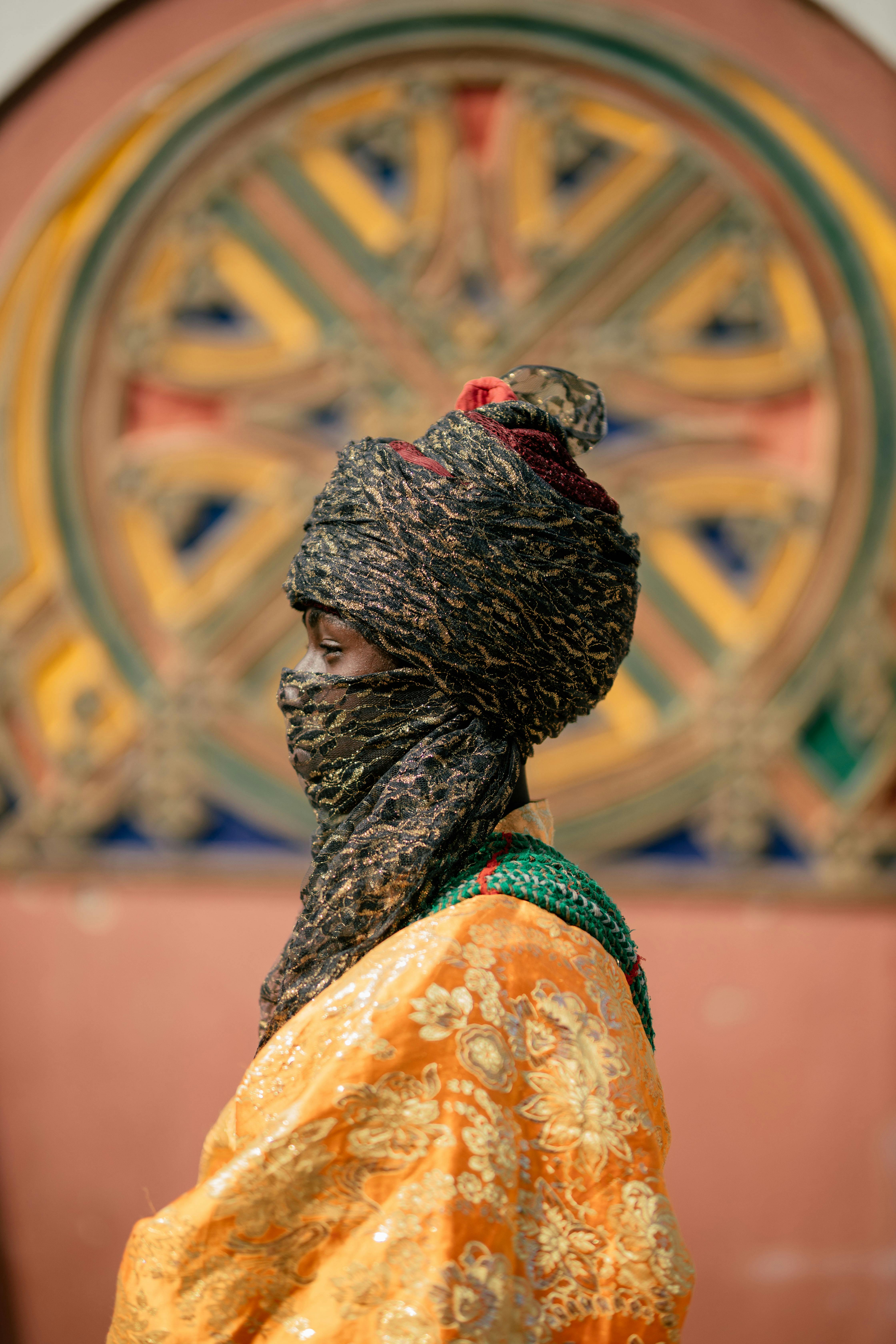 A person in vibrant traditional African clothing with intricate patterns and turban in front of a decorative backdrop.
