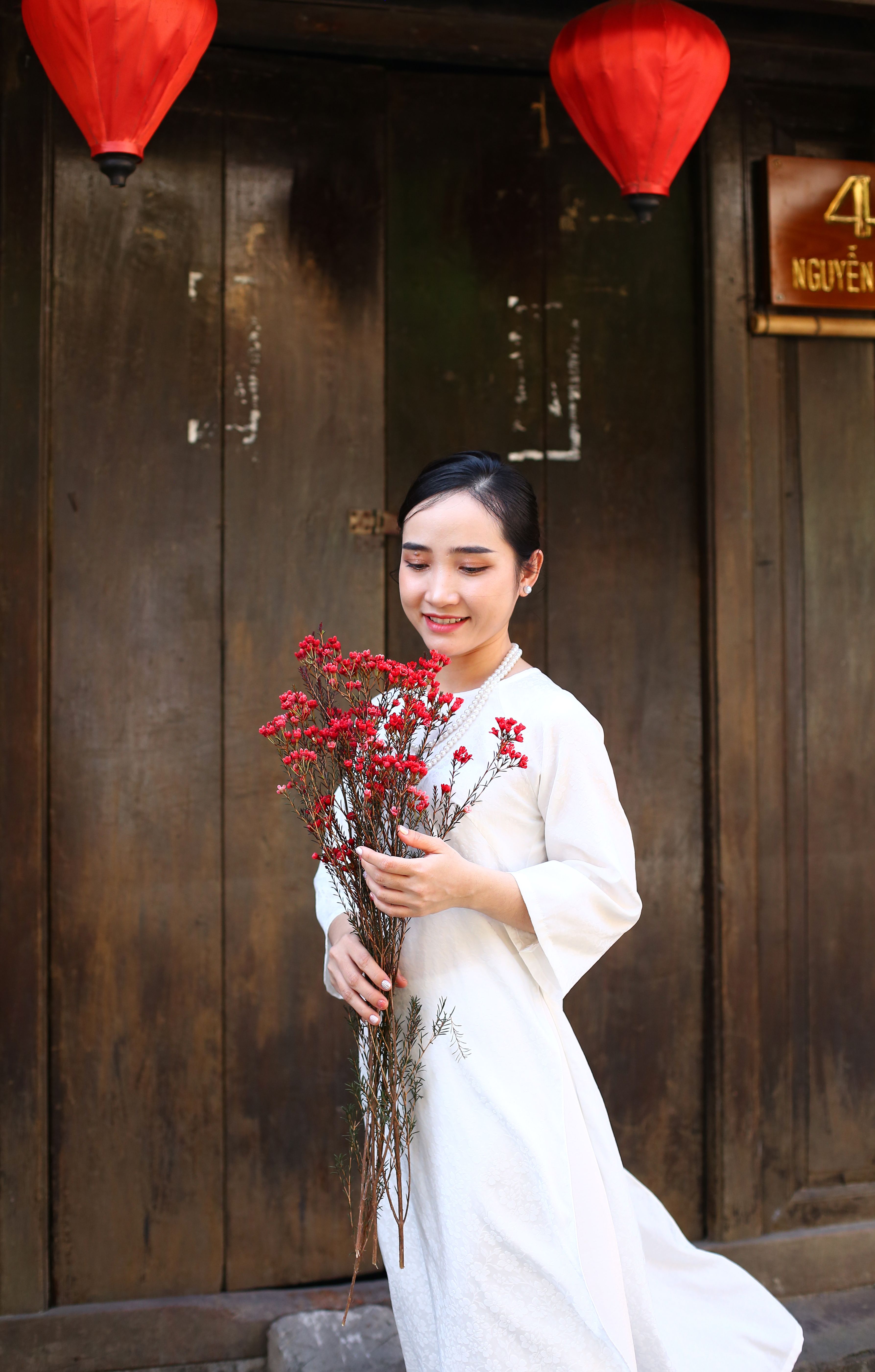 Smiling woman in traditional dress holding flowers in front of wooden doors in Hoi An, Vietnam.