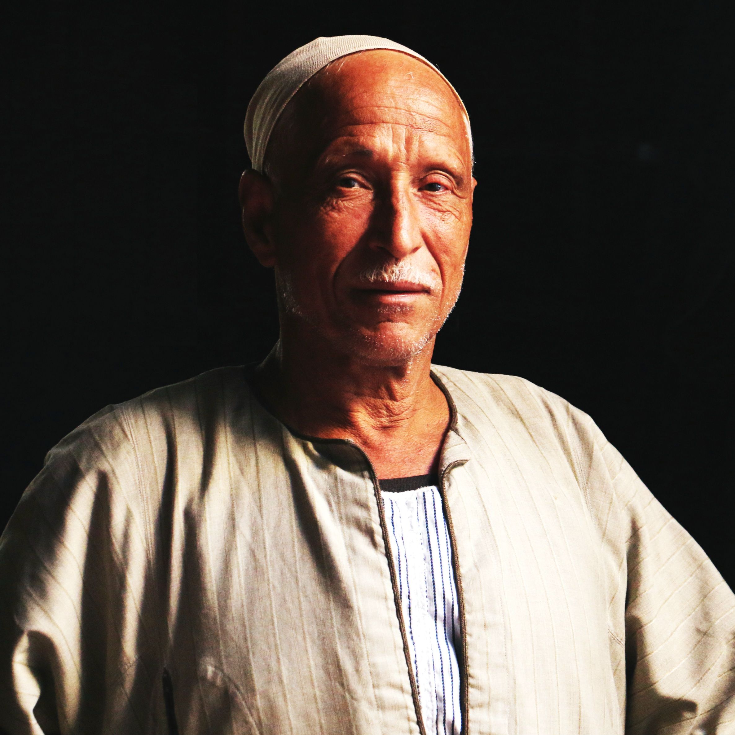 Elderly man wearing traditional clothing against a dark background.
