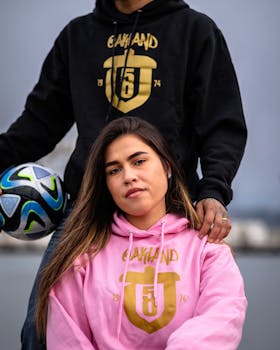 Two people wearing Oakland hoodies, posing with a soccer ball outdoors.