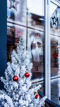 A festive snow-covered Christmas tree with red ornaments outside a shop window decorated for the holidays.
