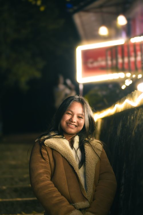 Free A joyful young woman in a warm jacket smiles on a well-lit night street. Stock Photo
