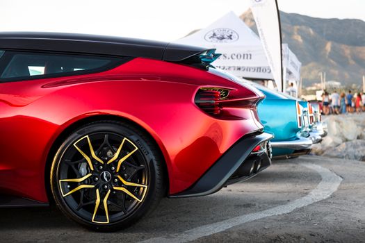 Sleek red sports car parked at a car show in Puerto Banus, Spain, with scenic mountain backdrop.