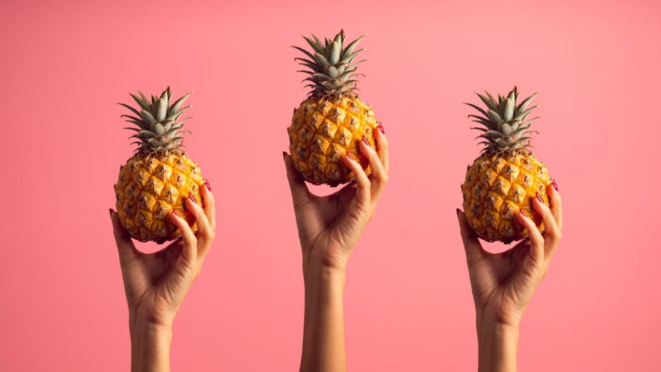 Three hands holding pineapples against a vibrant pink backdrop, emphasizing freshness and style.