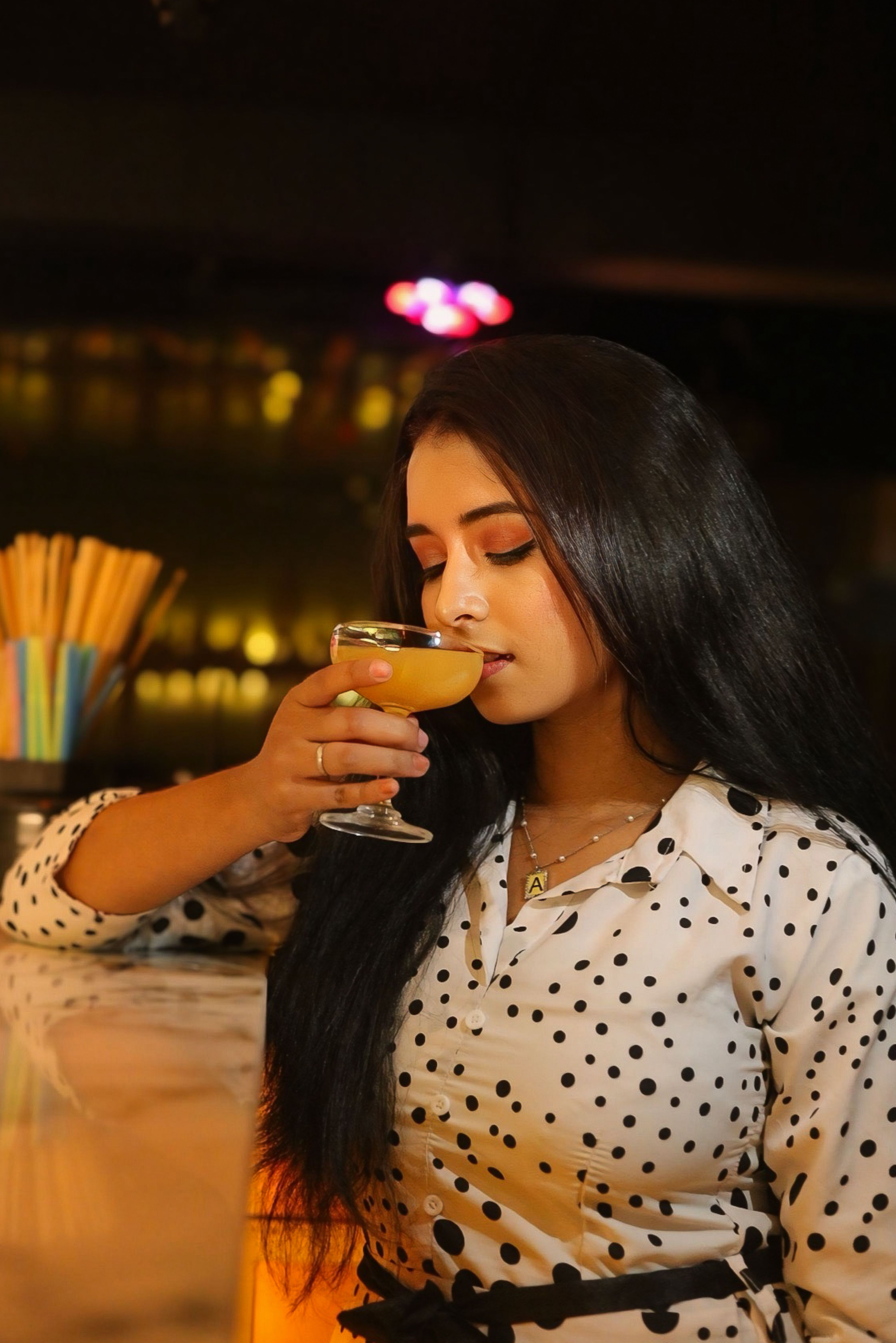 A woman in polka dot dress enjoys a cocktail in a dimly lit cafe.