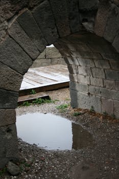Stone archway framing a reflective puddle on a wet surface, outdoors.