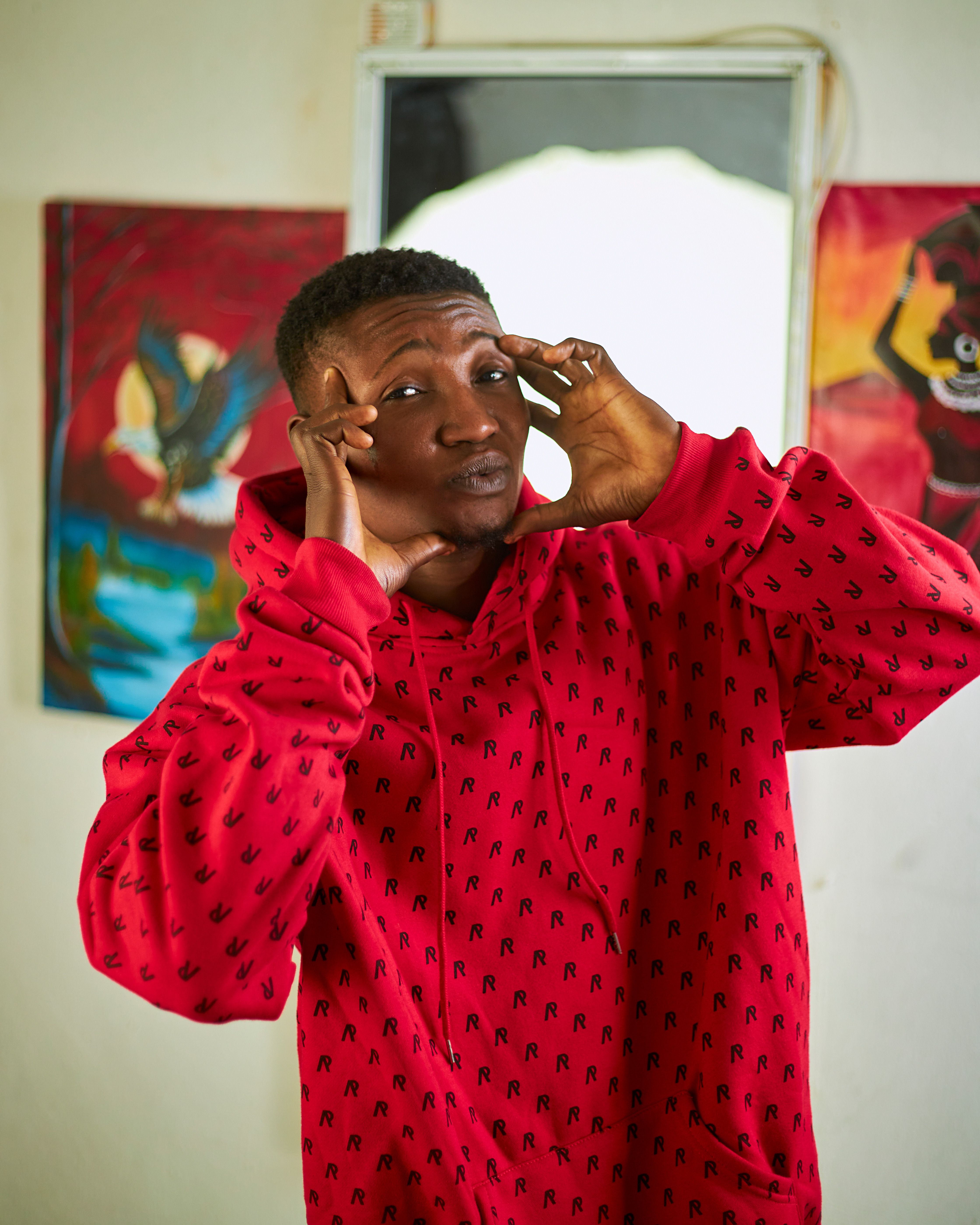 A young man making a playful expression while wearing a red hoodie, indoor setting with colorful art in the background.