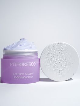 Purple jar of soothing cream with lid droplets under bright lighting. Ideal for skincare and beauty concepts.
