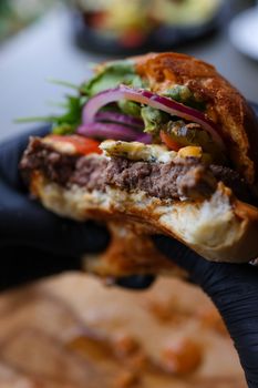 Close-up of a juicy half-eaten burger with fresh toppings held in a hand with gloves.