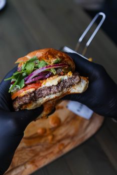 Juicy hamburger with fresh toppings held by gloved hands on wooden board.