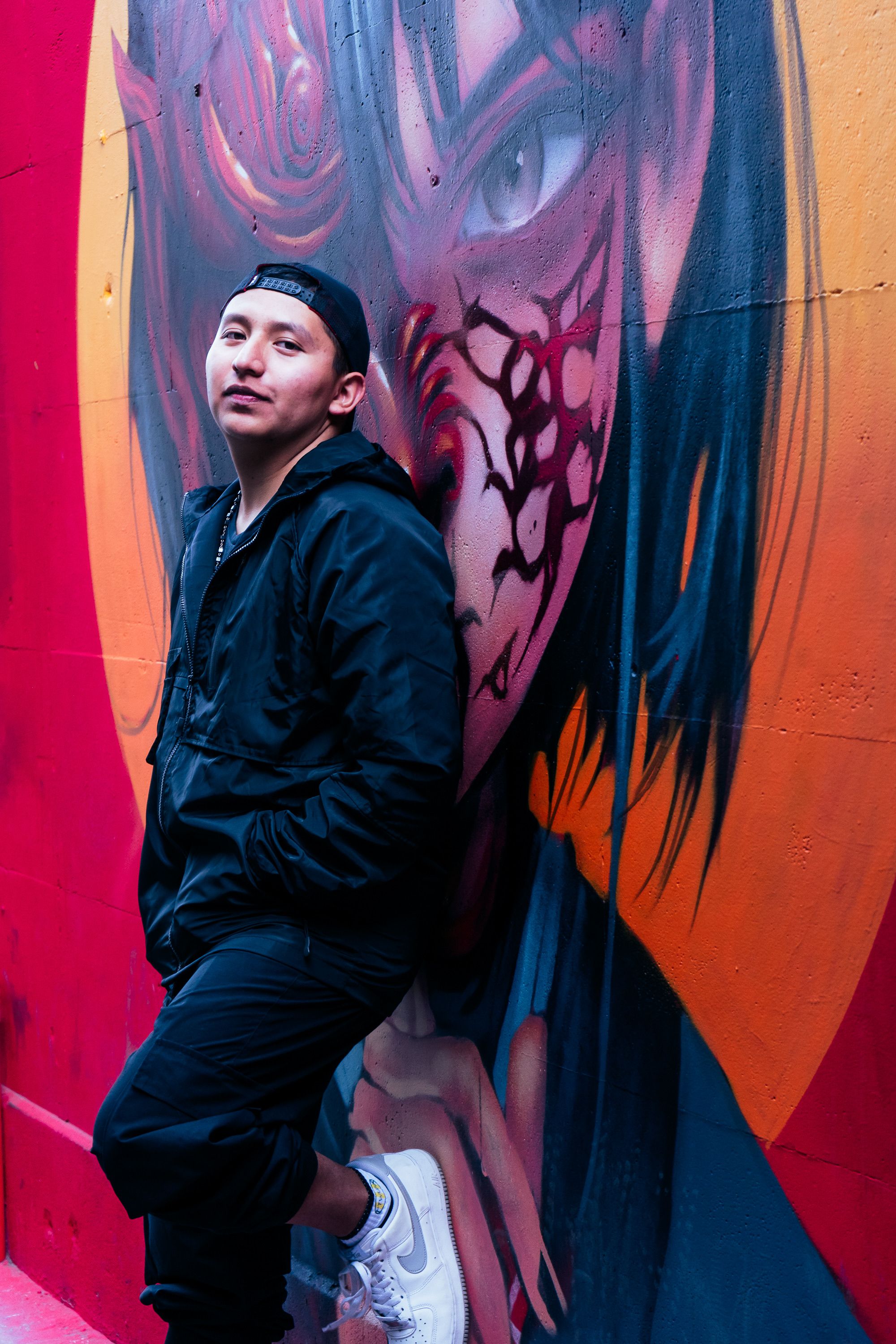 Young man leans against colorful street art mural in an urban alleyway.
