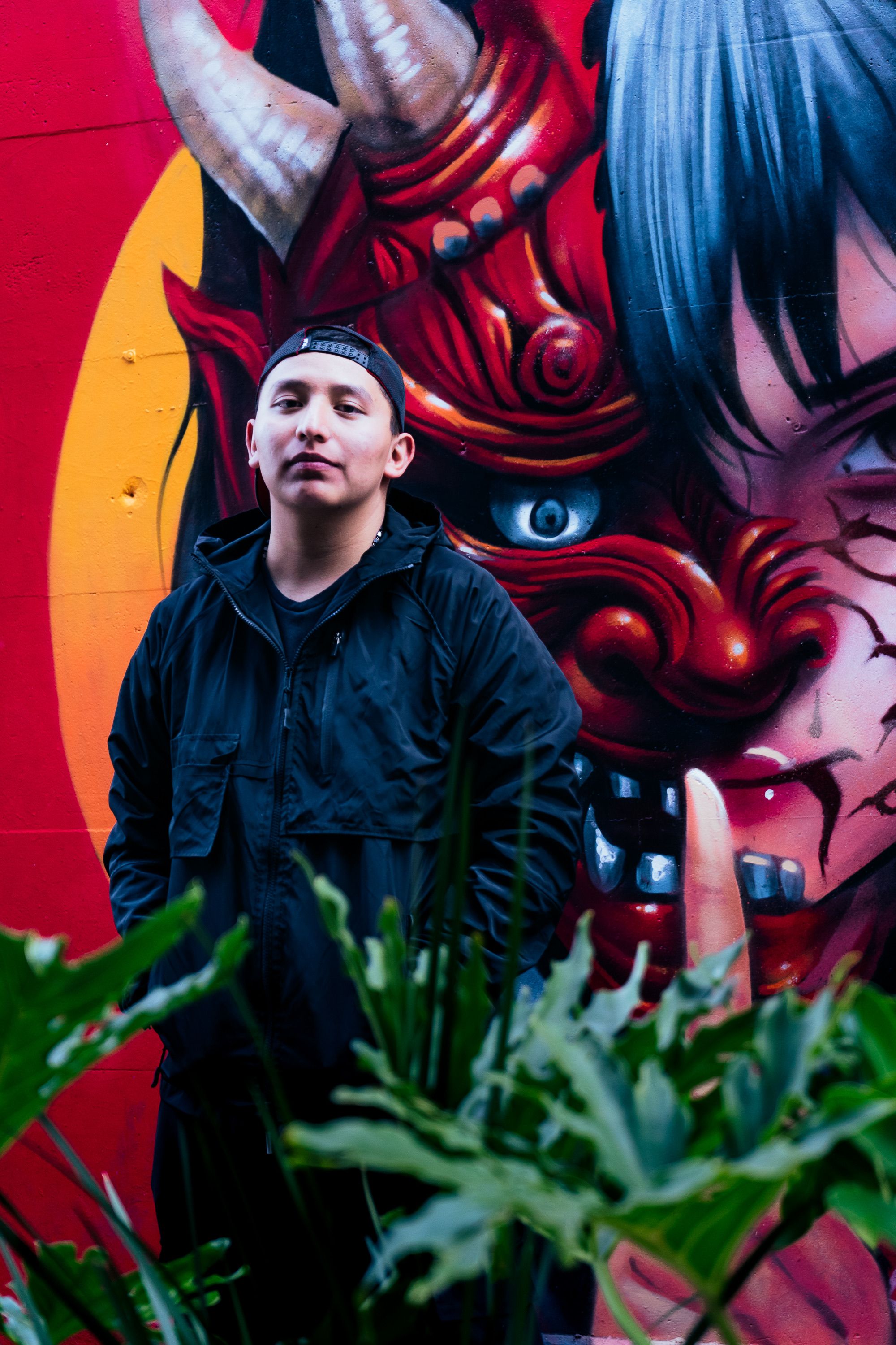 Man posing in front of colorful graffiti mural featuring a traditional Japanese oni mask.