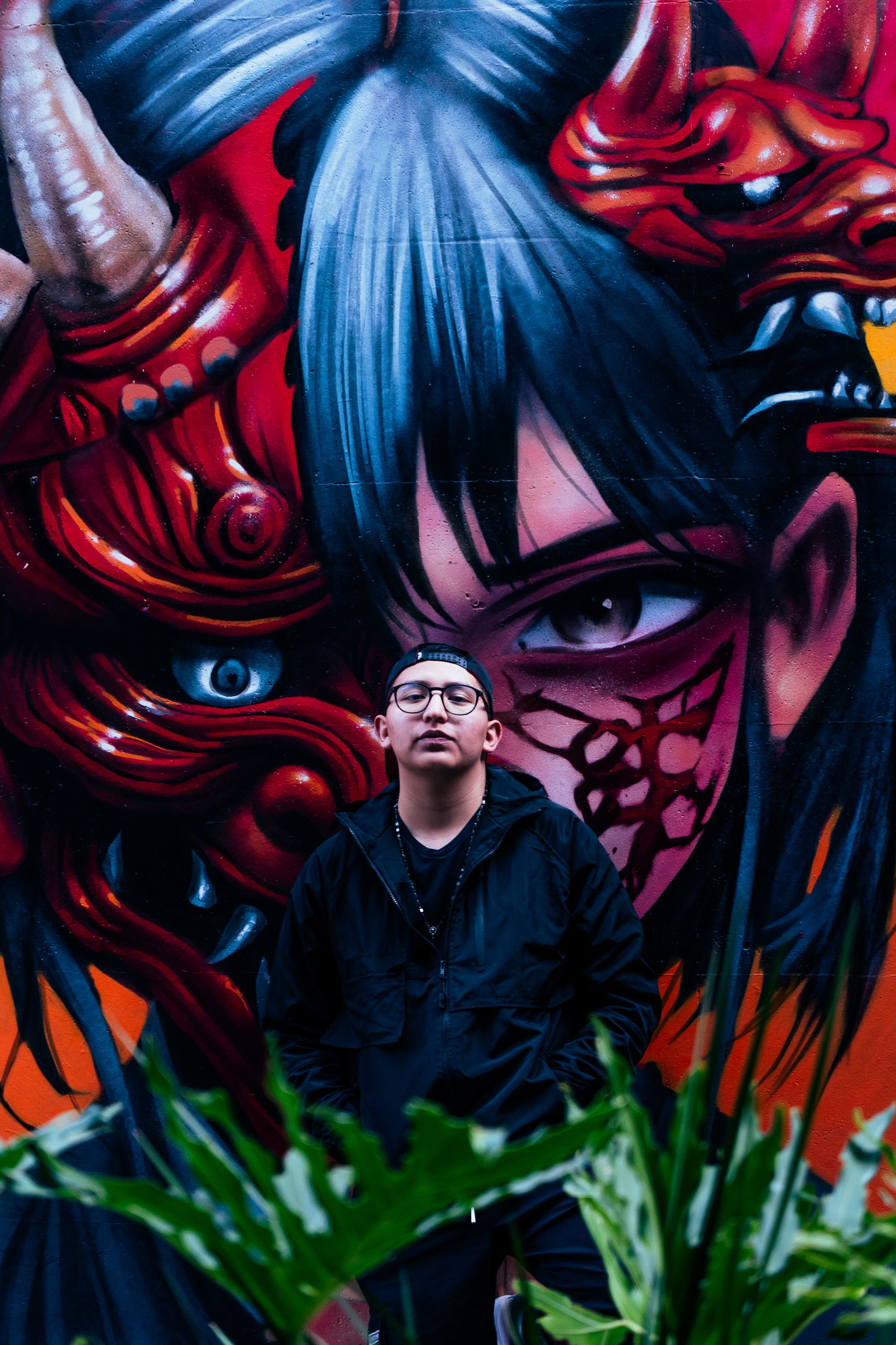 Young man in front of colorful street art depicting a fantasy character with demon mask.