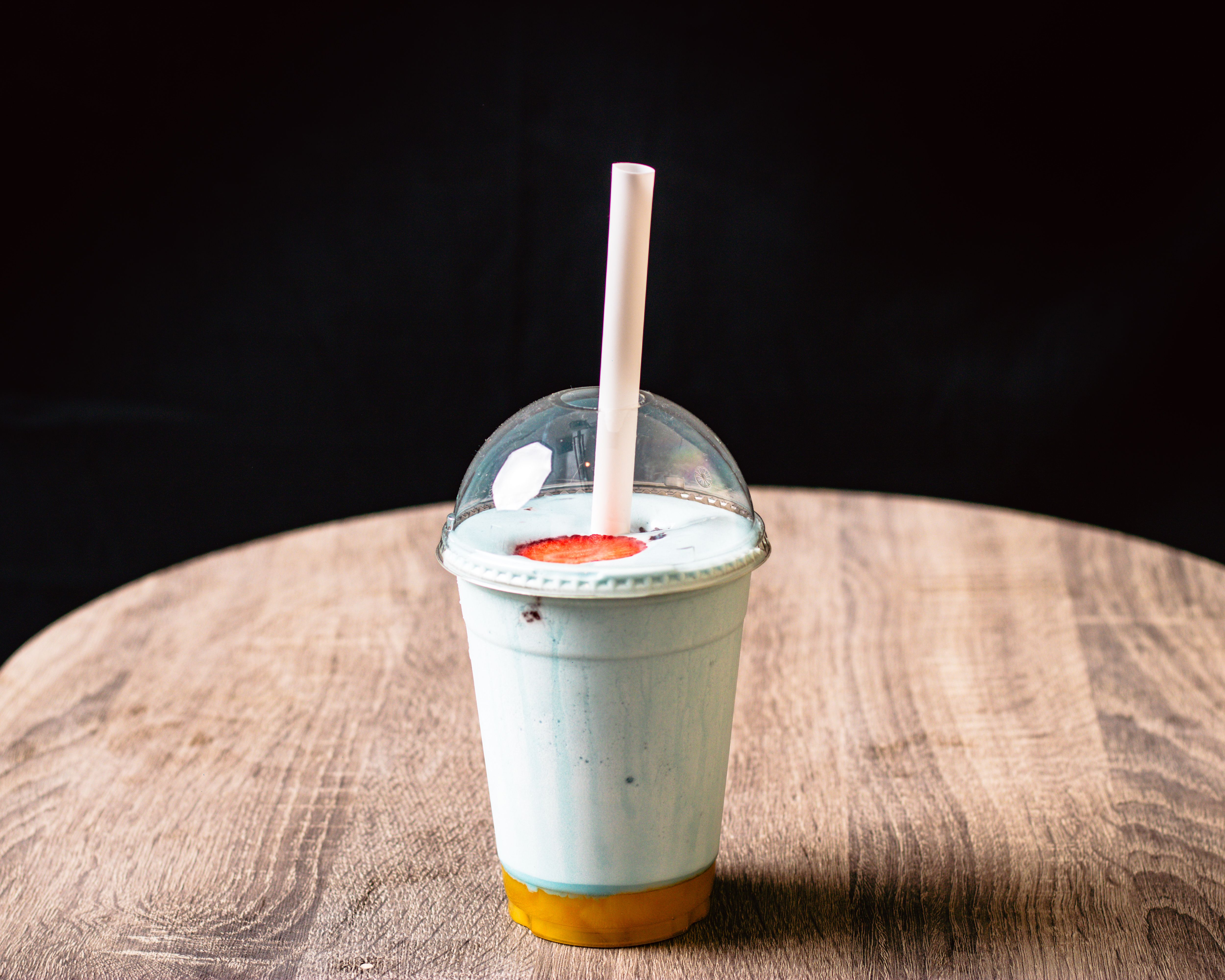 A blue milkshake in a plastic cup with dome lid and straw on a wooden table.