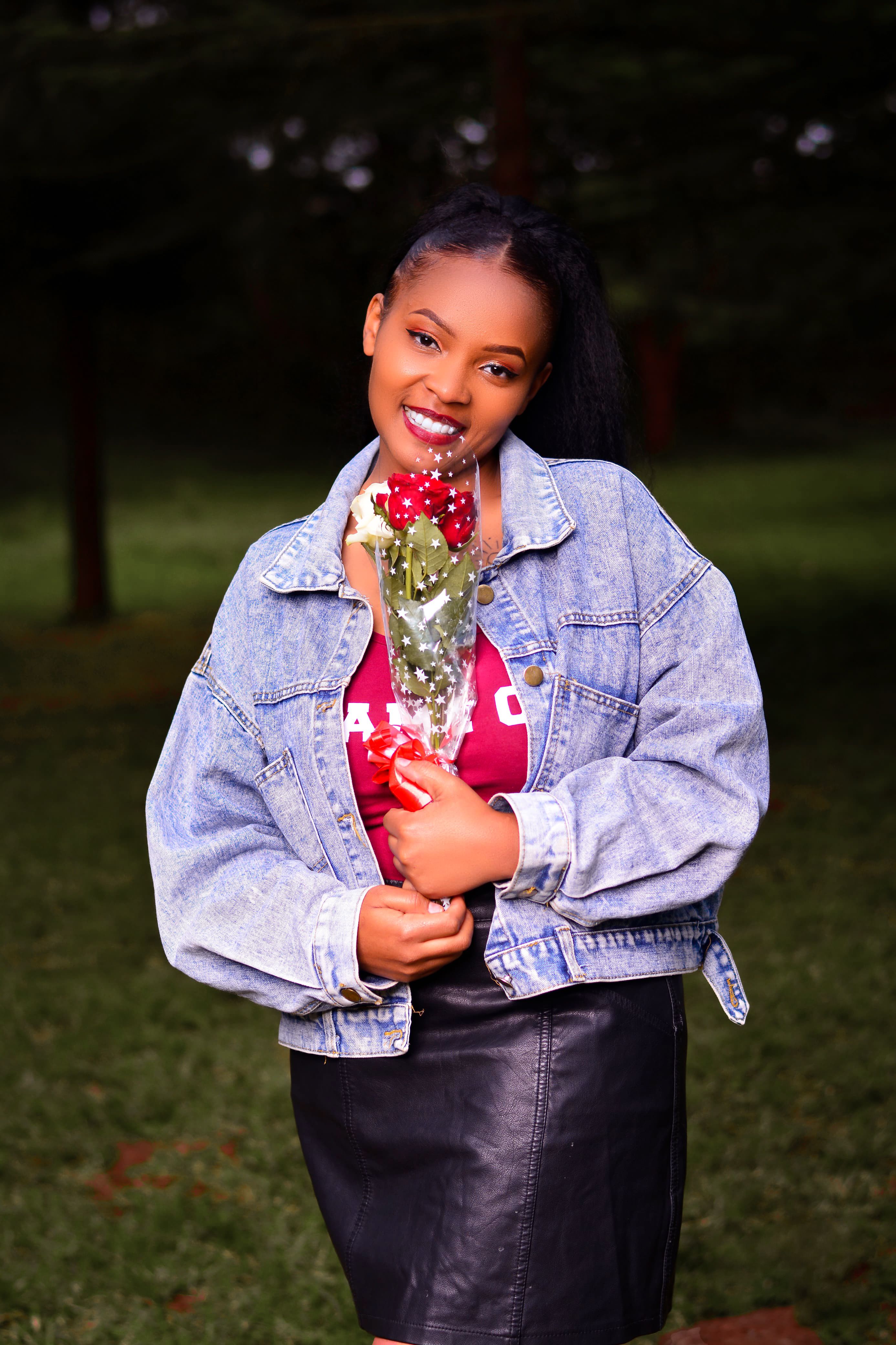 A woman in a denim jacket holding a bouquet of roses while smiling in a park.