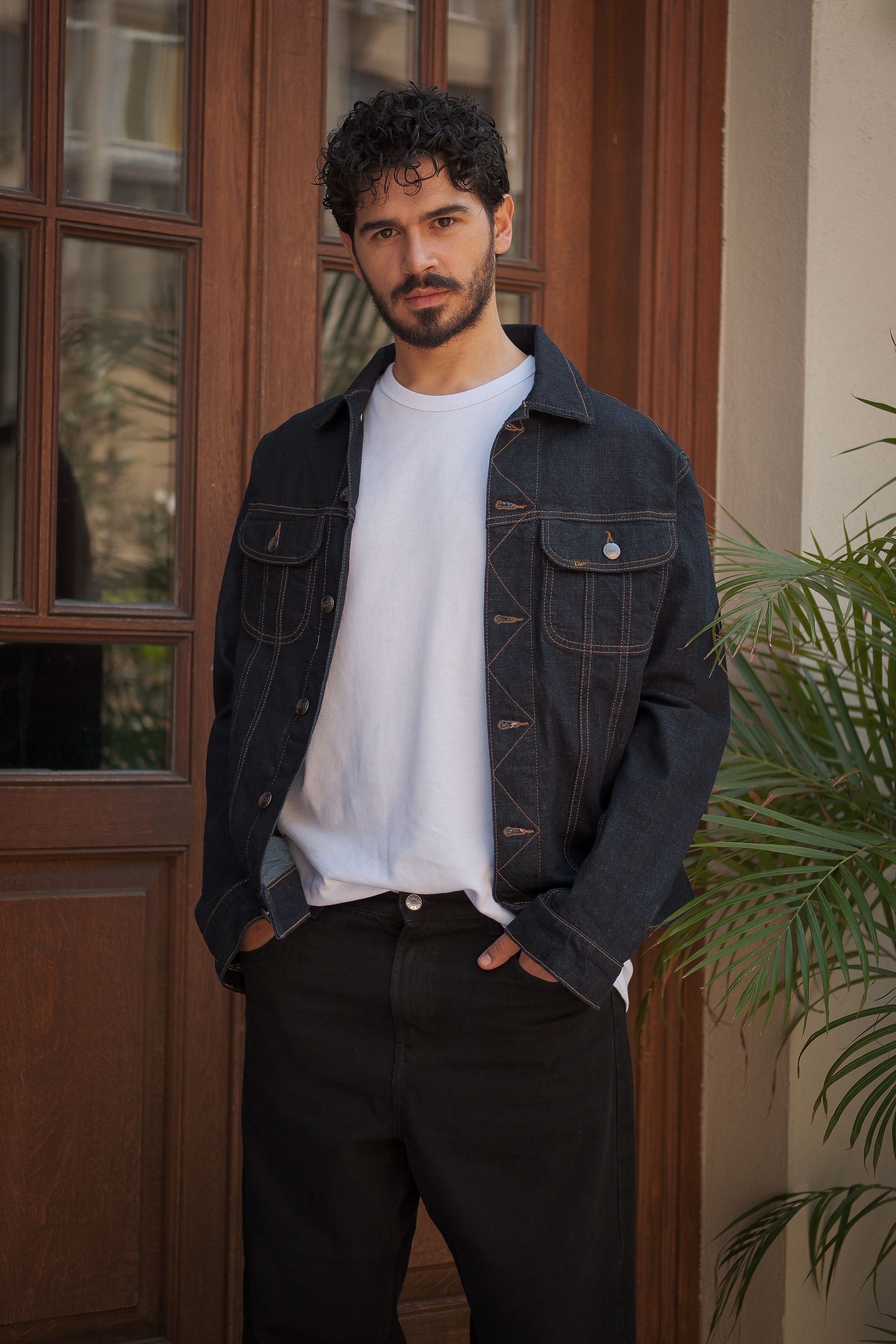 Man wearing denim jacket and white t-shirt poses confidently outdoors.