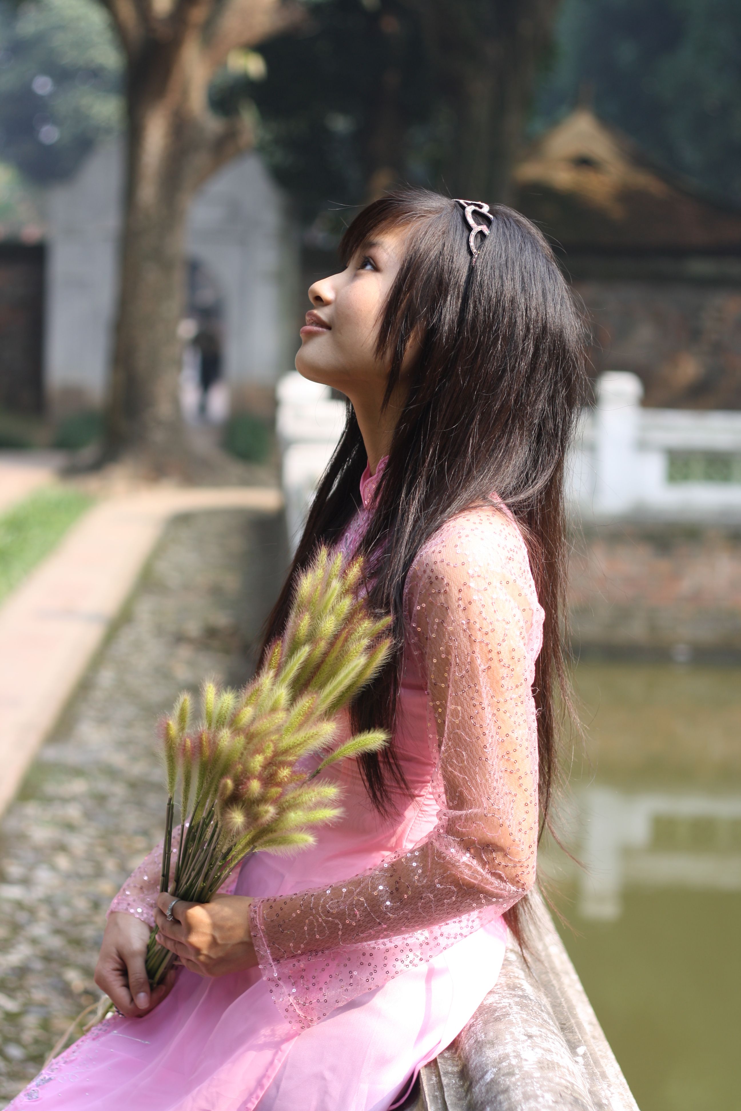 Young woman in pink dress holding flowers, looking upwards, outdoors.