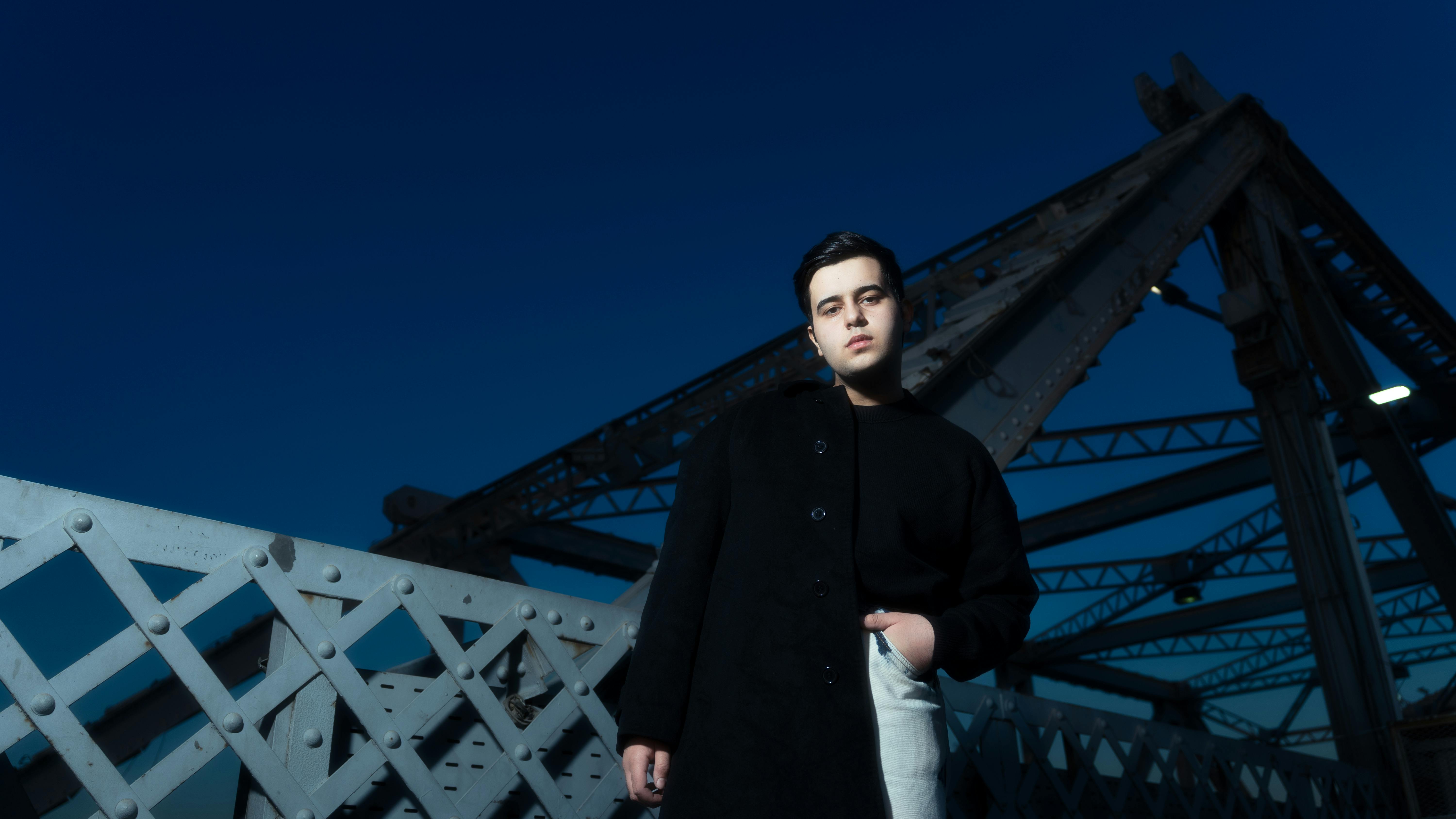 Young man in black coat posing on an industrial bridge at dusk.