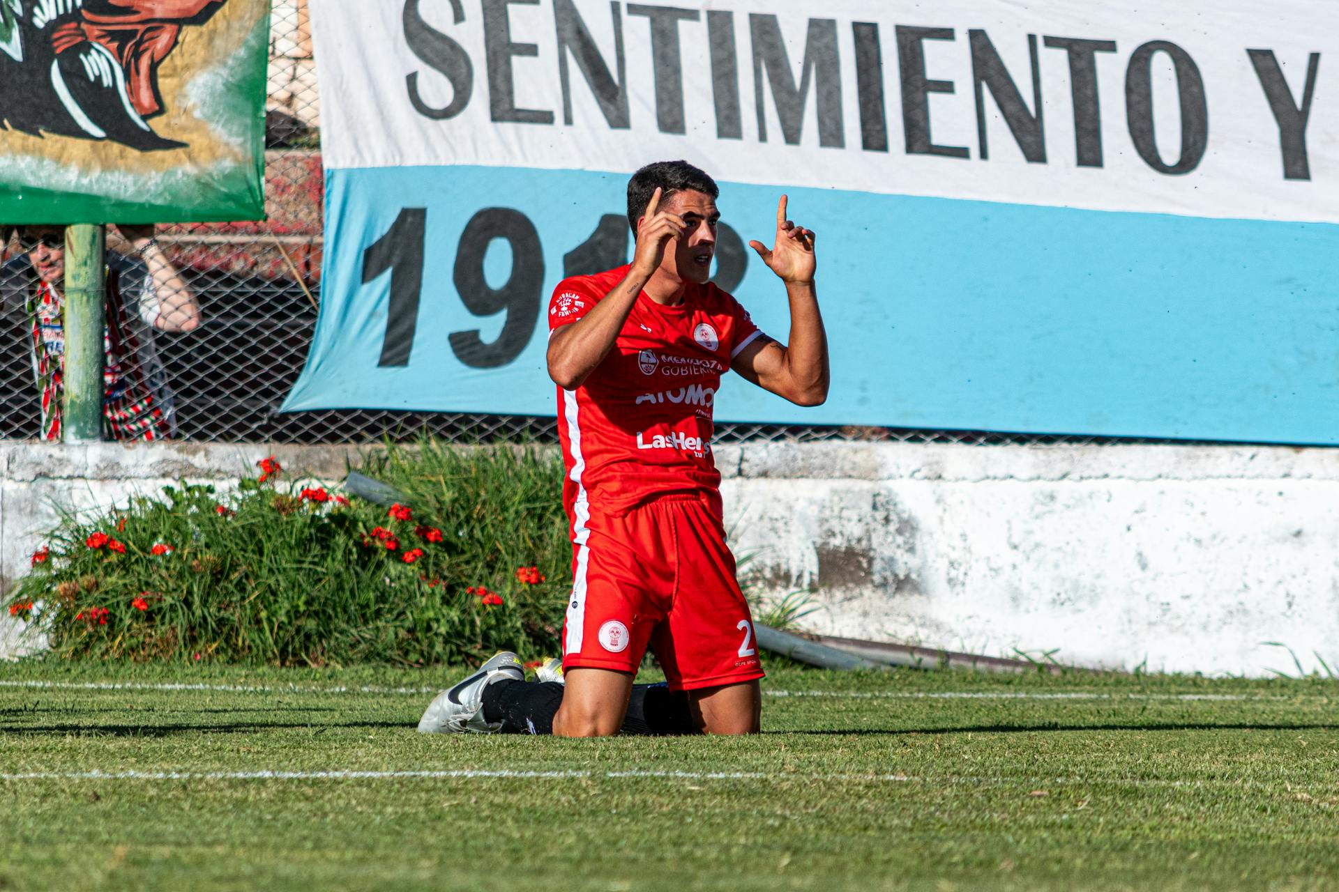 A soccer player in red kit celebrates after scoring on a sunny day.