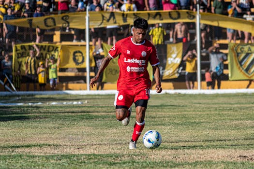 A soccer player in red uniform dribbling during a daytime match with spectators.