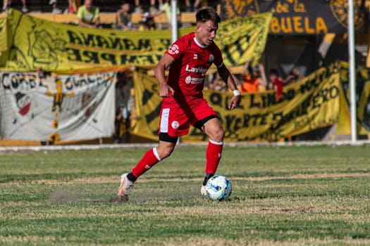 Soccer player in red kit dribbling the ball during a daytime match.