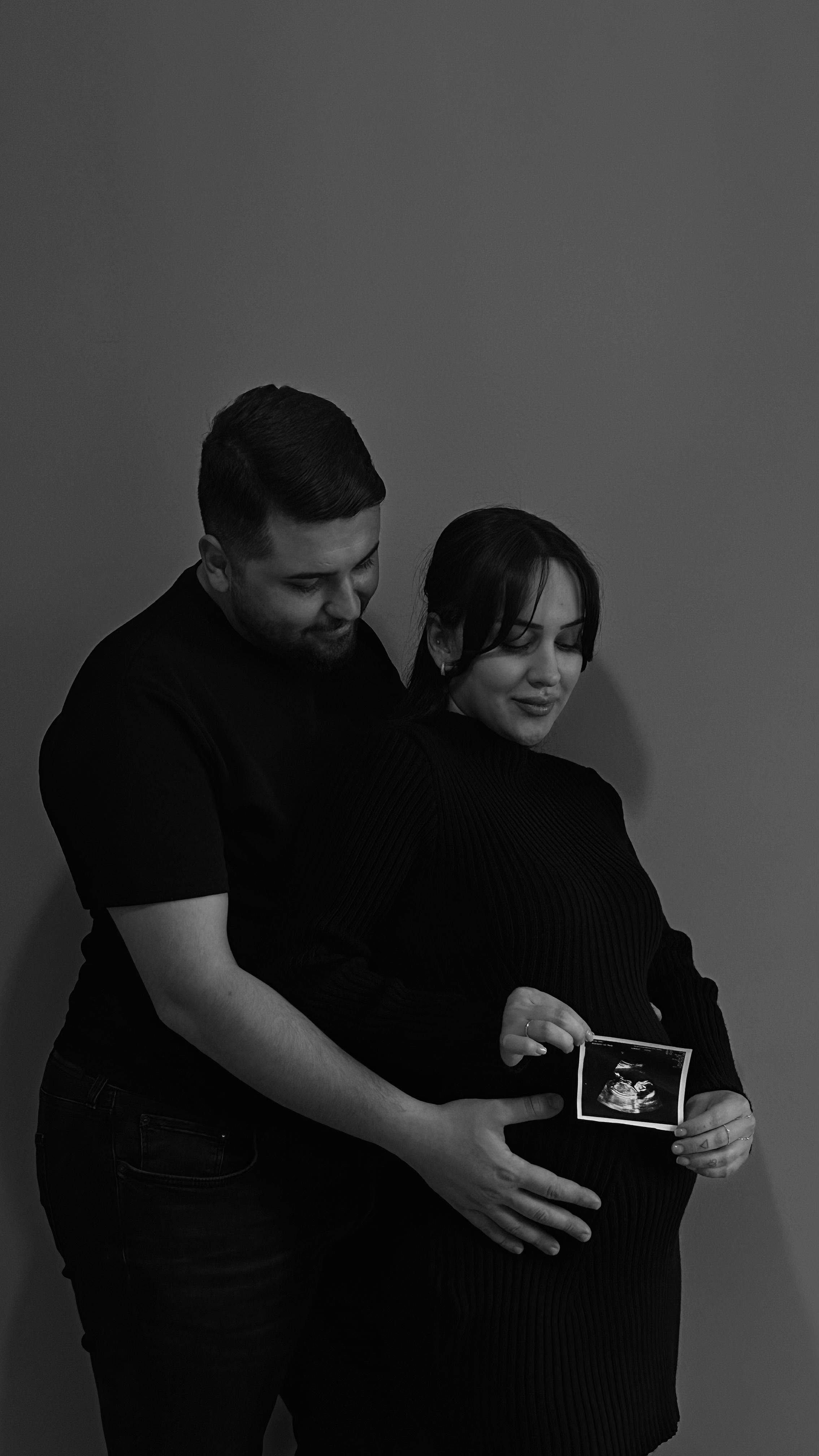 Expecting couple in black attire holding ultrasound photo, celebrating pregnancy.