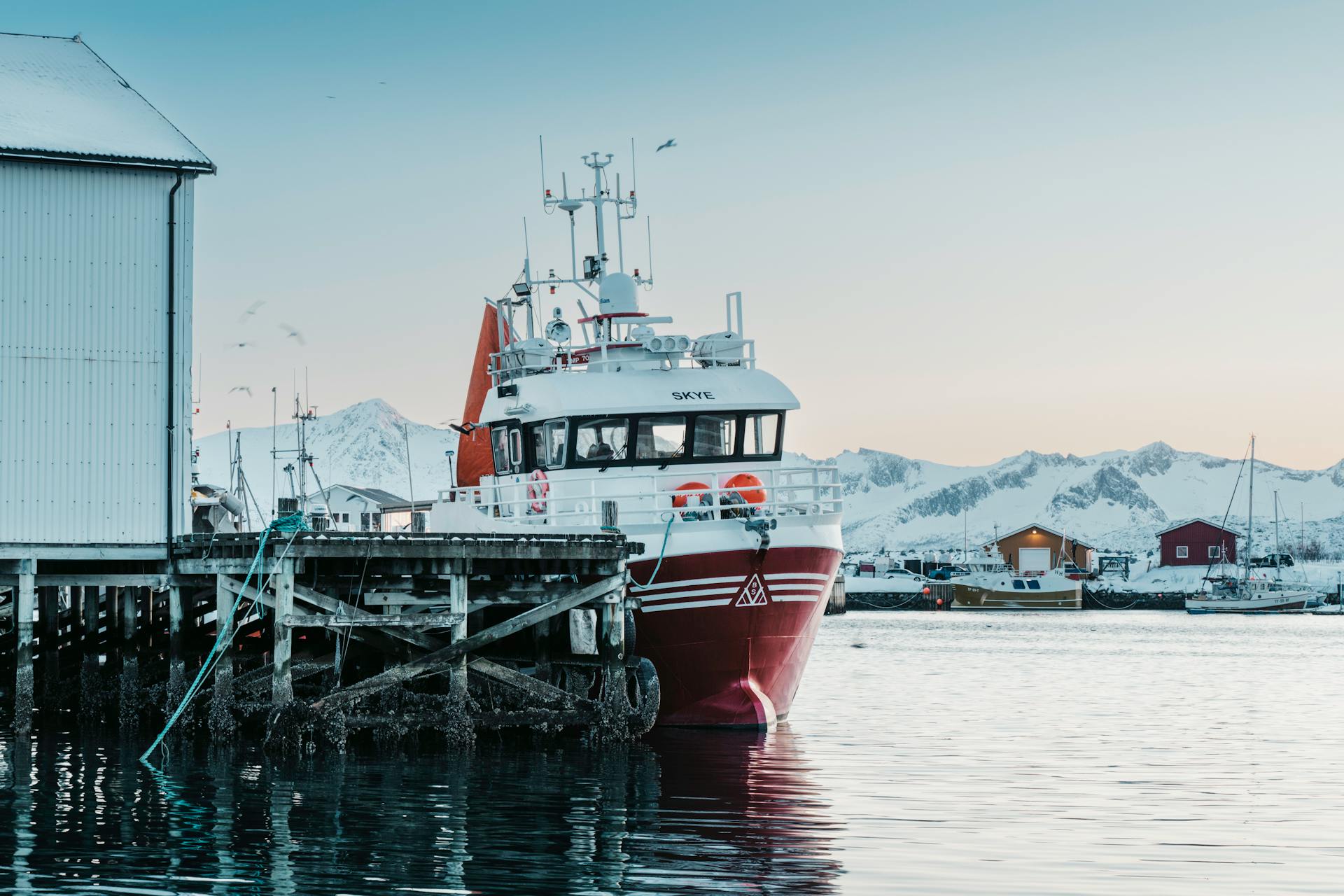 A vibrant fishing boat docked at a snowy harbor in Norway with mountains in the background.