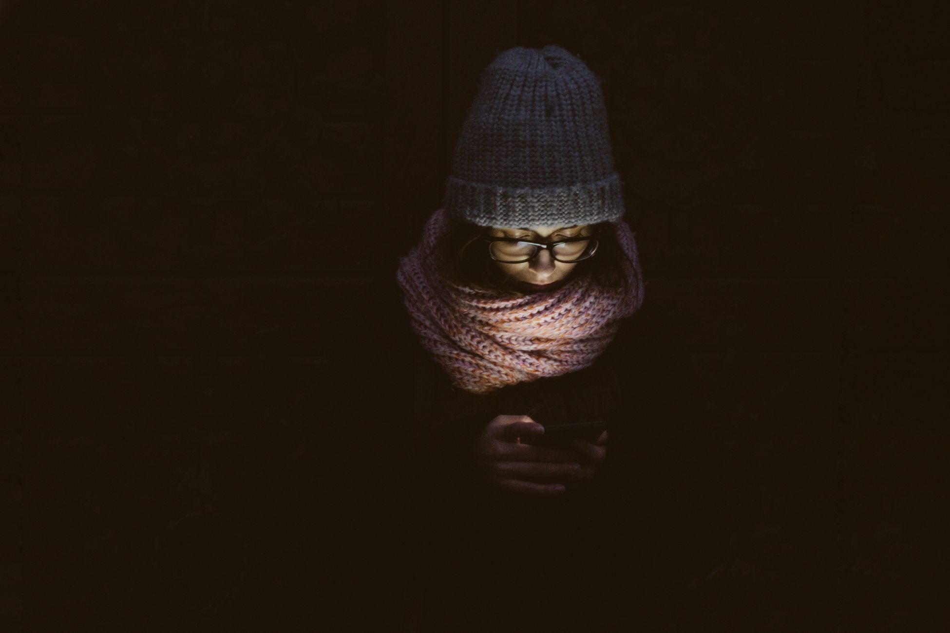 A woman with a hat and scarf reading her smartphone in a dark setting, softly lit.