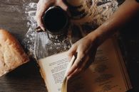 Hands with coffee cup and recipe book on flour-dusted table with bread.