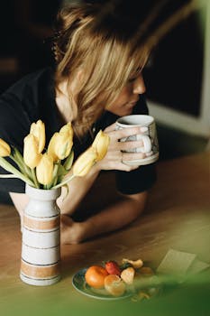 Woman sipping coffee surrounded by tulips and fruits on a sunny morning indoors.