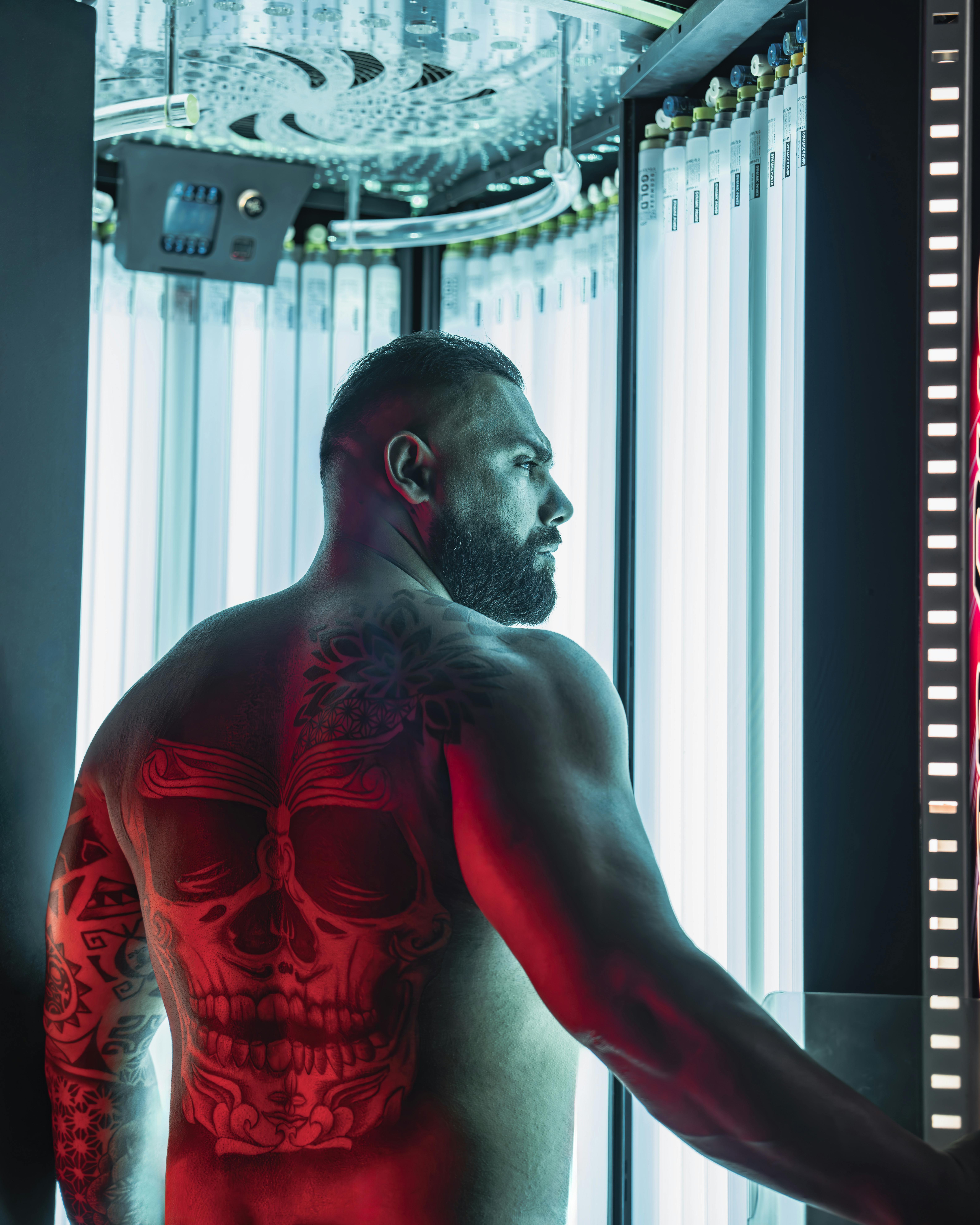 Muscular man with skull tattoo using a vertical tanning booth, lit with intense and dramatic lighting.