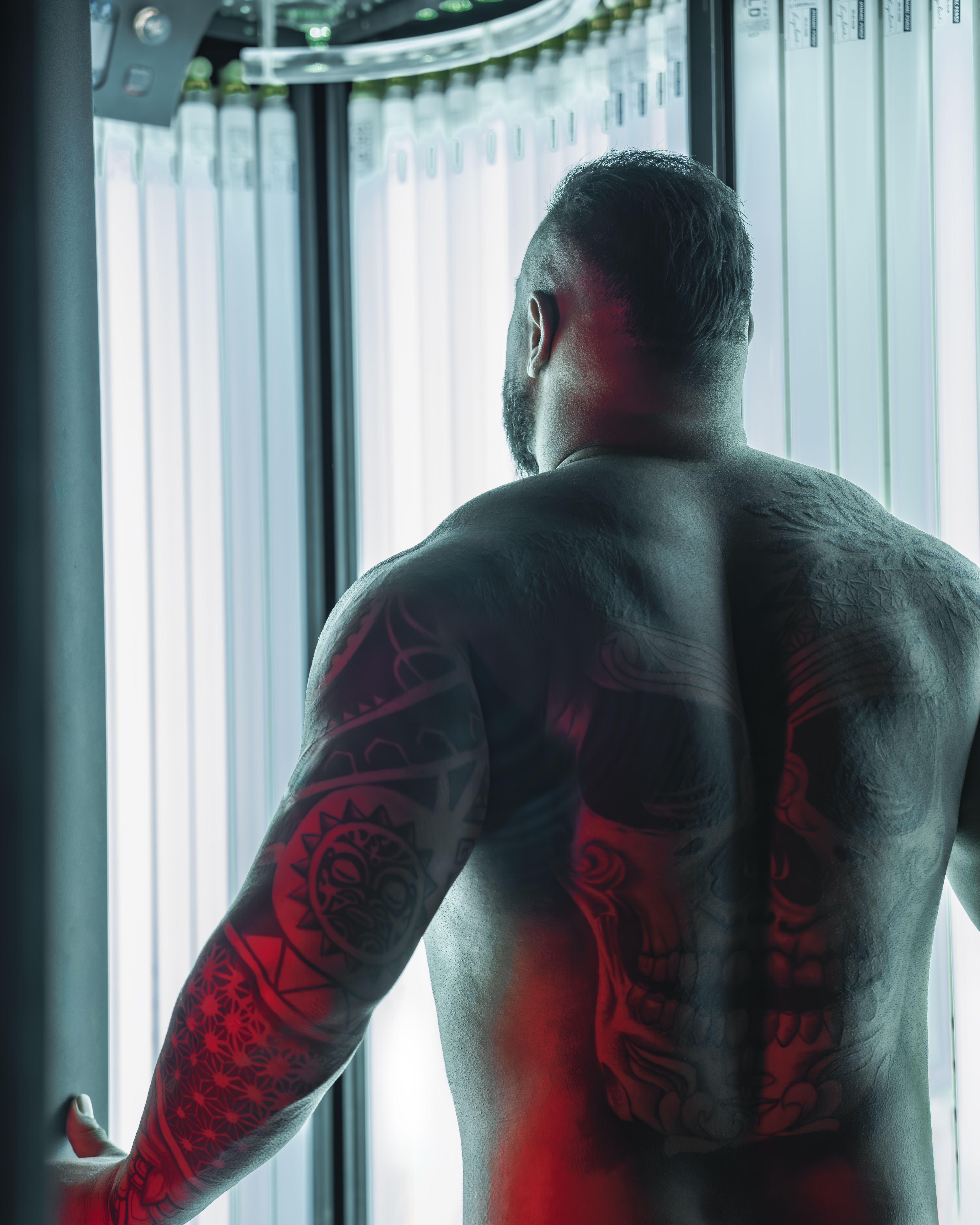 A muscular tattooed man stands in a tanning booth seen from behind, showcasing his back tattoos.