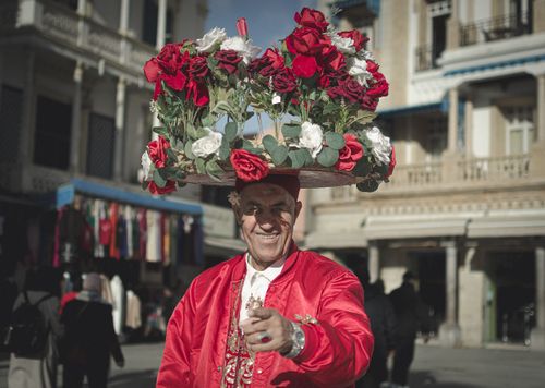 Free An elderly flower vendor in vibrant attire smiles while balancing a tray of roses in a Tunisian market. Stock Photo