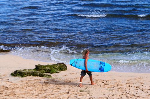 Free A surfer walks along Hana Beach in Hawaii carrying a vibrant blue surfboard near the waves. Stock Photo