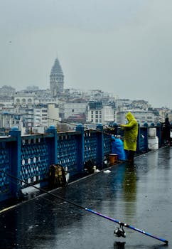 A fisherman in a yellow raincoat casts a line on the wet Galata Bridge with the iconic Galata Tower in the background in Istanbul.