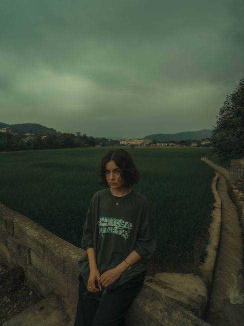 A thoughtful woman stands in a dark green field under a cloudy sky, suggesting a moody atmosphere.