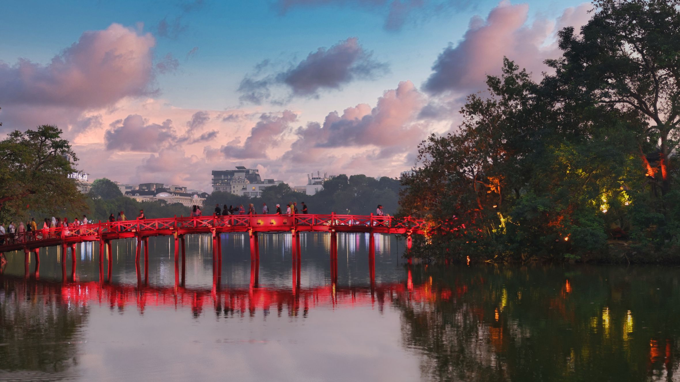 Scenic view of the Huc Bridge in Hanoi, Vietnam, illuminated at twilight over Hoan Kiem Lake with reflections and vibrant sky.