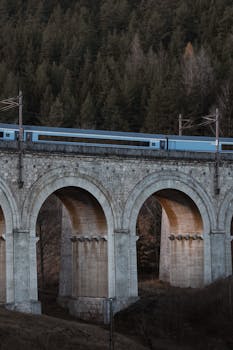 A blue train travels over the historic Semmering Railway viaduct amidst a lush forest in Austria.