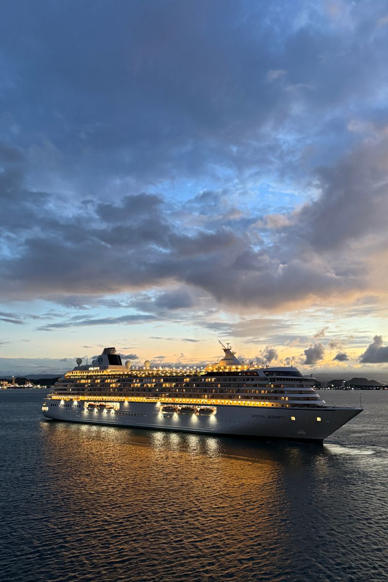 A majestic cruise ship sails on the open sea under a dramatic sunset sky, reflecting golden hues.