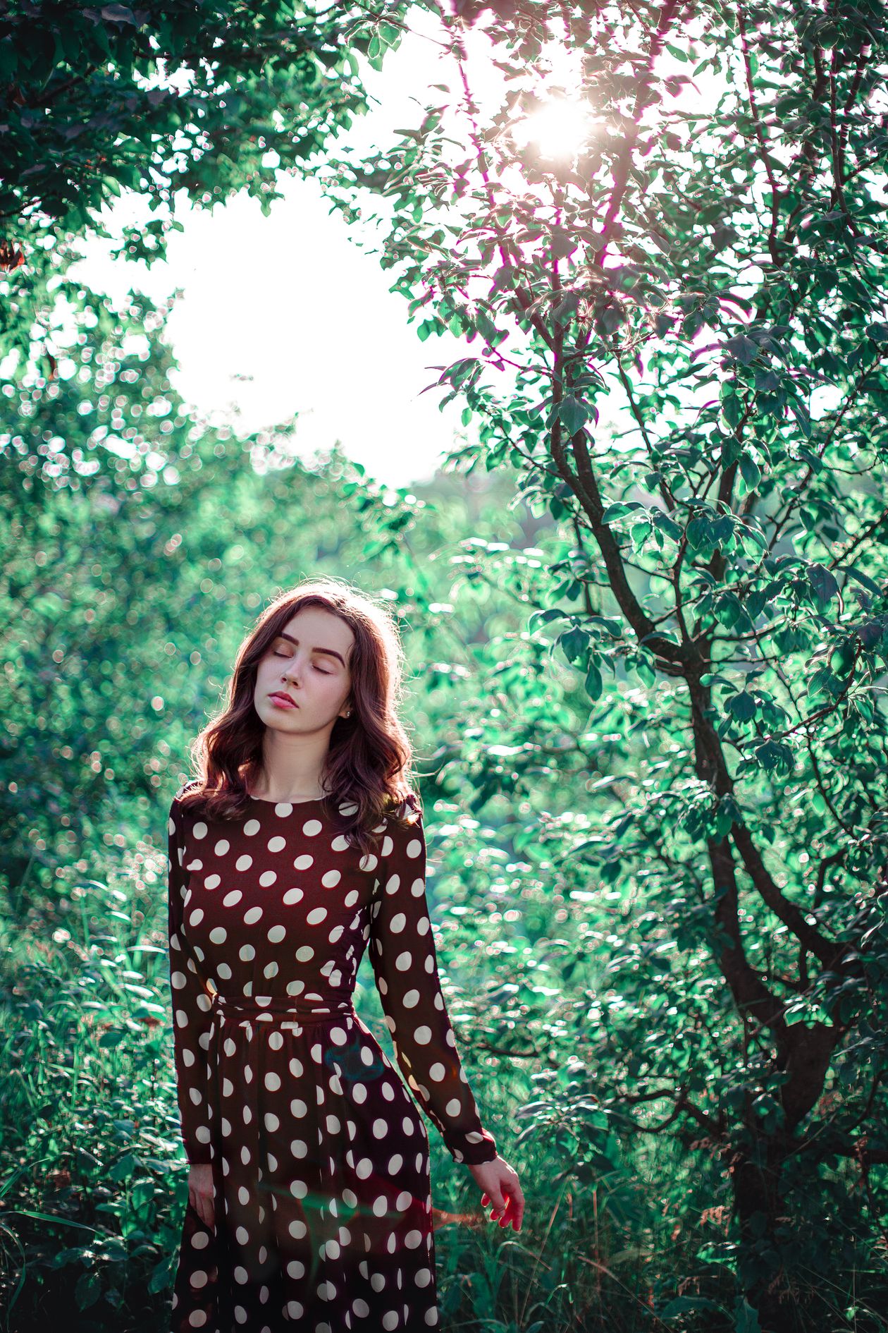 Woman in a brown polka-dot dress standing in a sunlit garden