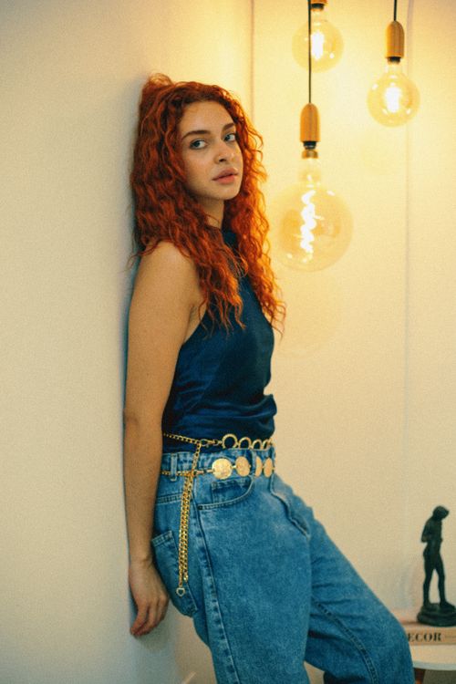 Free Fashionable woman with curly hair stands against wall with hanging bulbs in São Paulo. Stock Photo