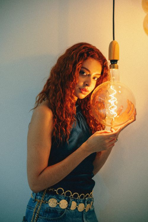 Free A young woman with curly hair holds a glowing bulb, creating a warm ambiance. Stock Photo