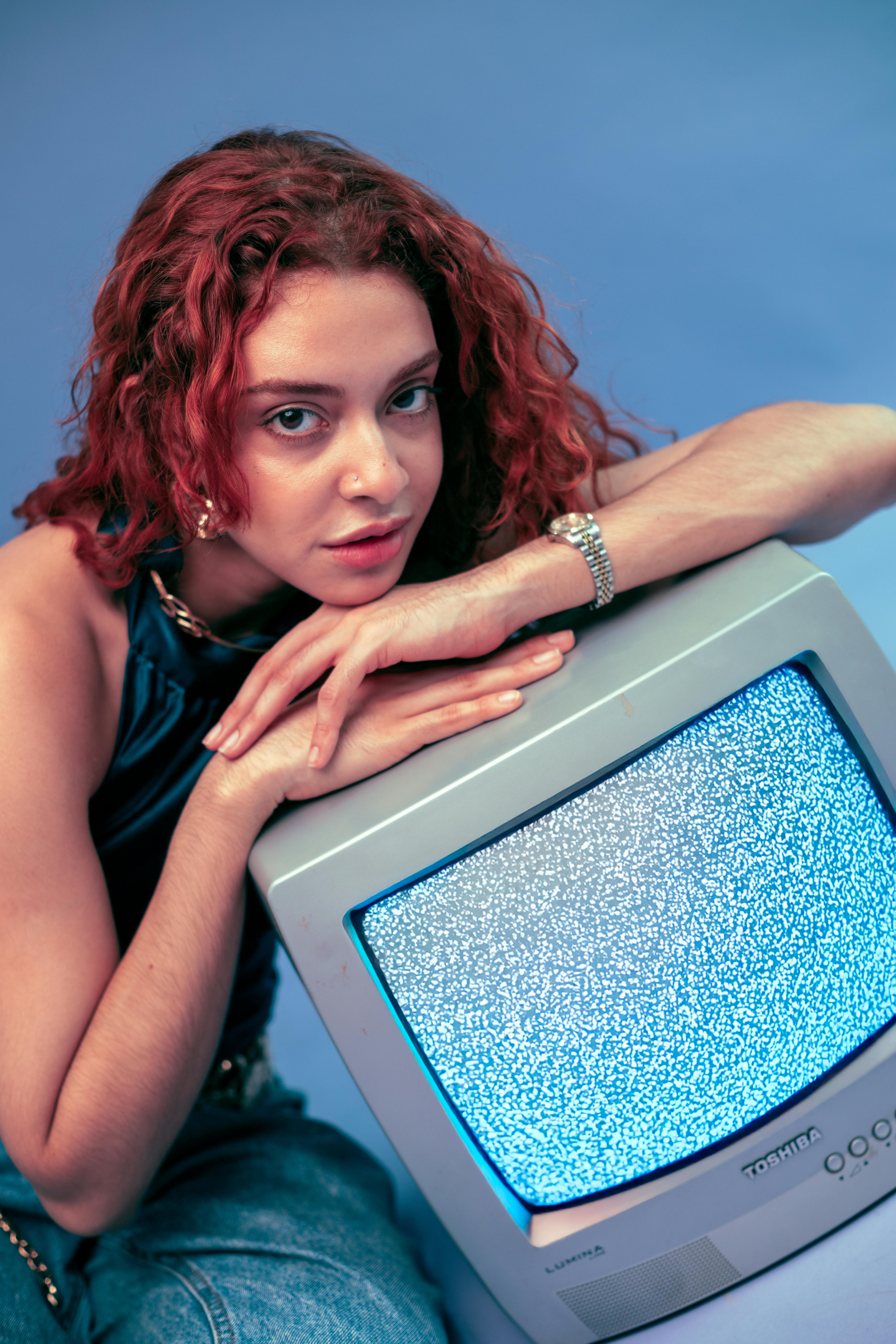 Portrait of a young woman with curly red hair leaning on an old TV showing static.
