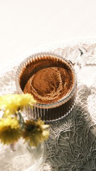 Close-up of a coffee cup with foam on a lace tablecloth, accompanied by yellow flowers.