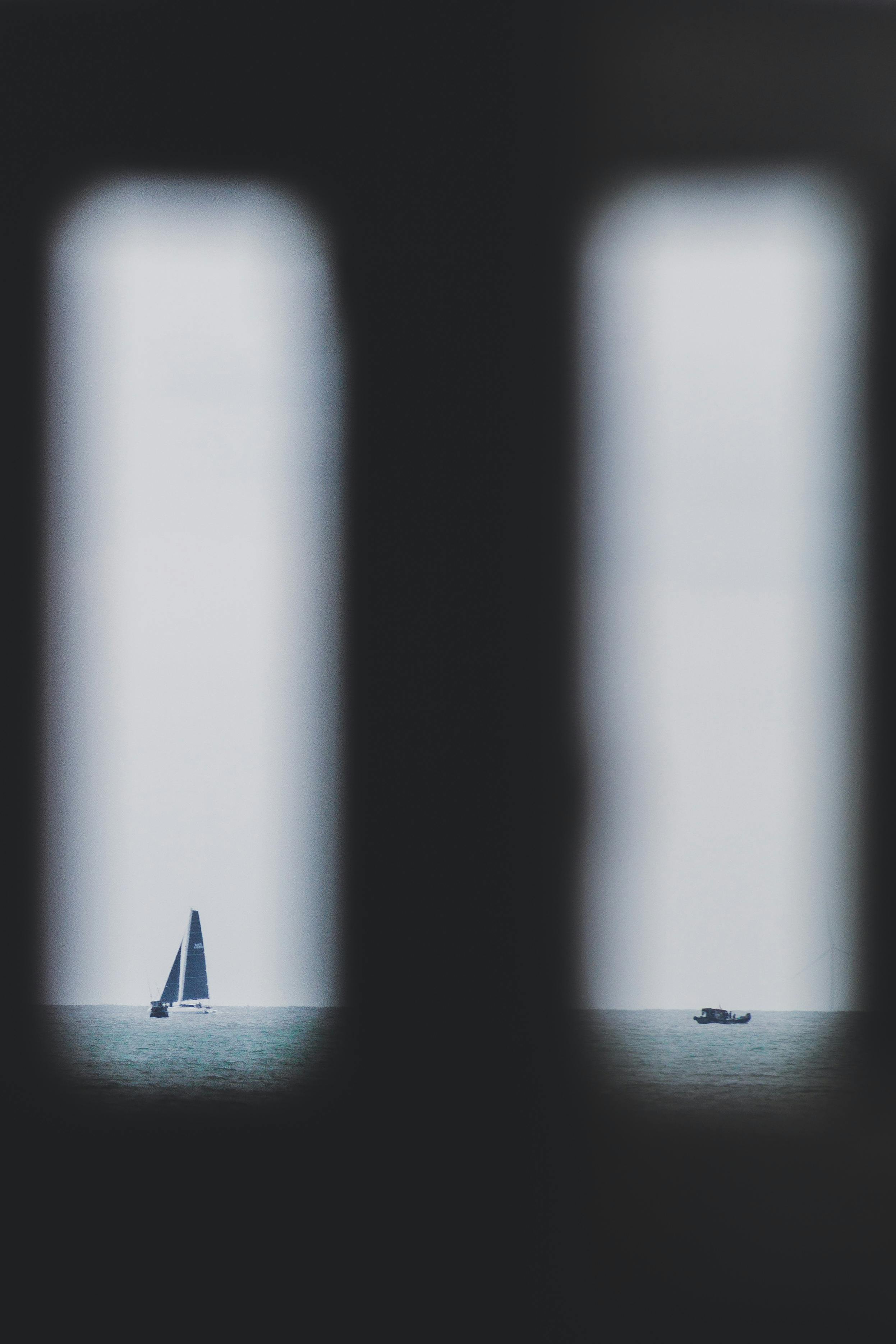 Free A sailboat and a distant boat seen through a window frame with a moody atmosphere. Stock Photo