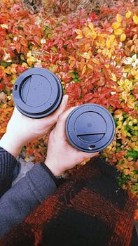 Two people holding coffee cups surrounded by vibrant autumn leaves, perfect fall vibe.