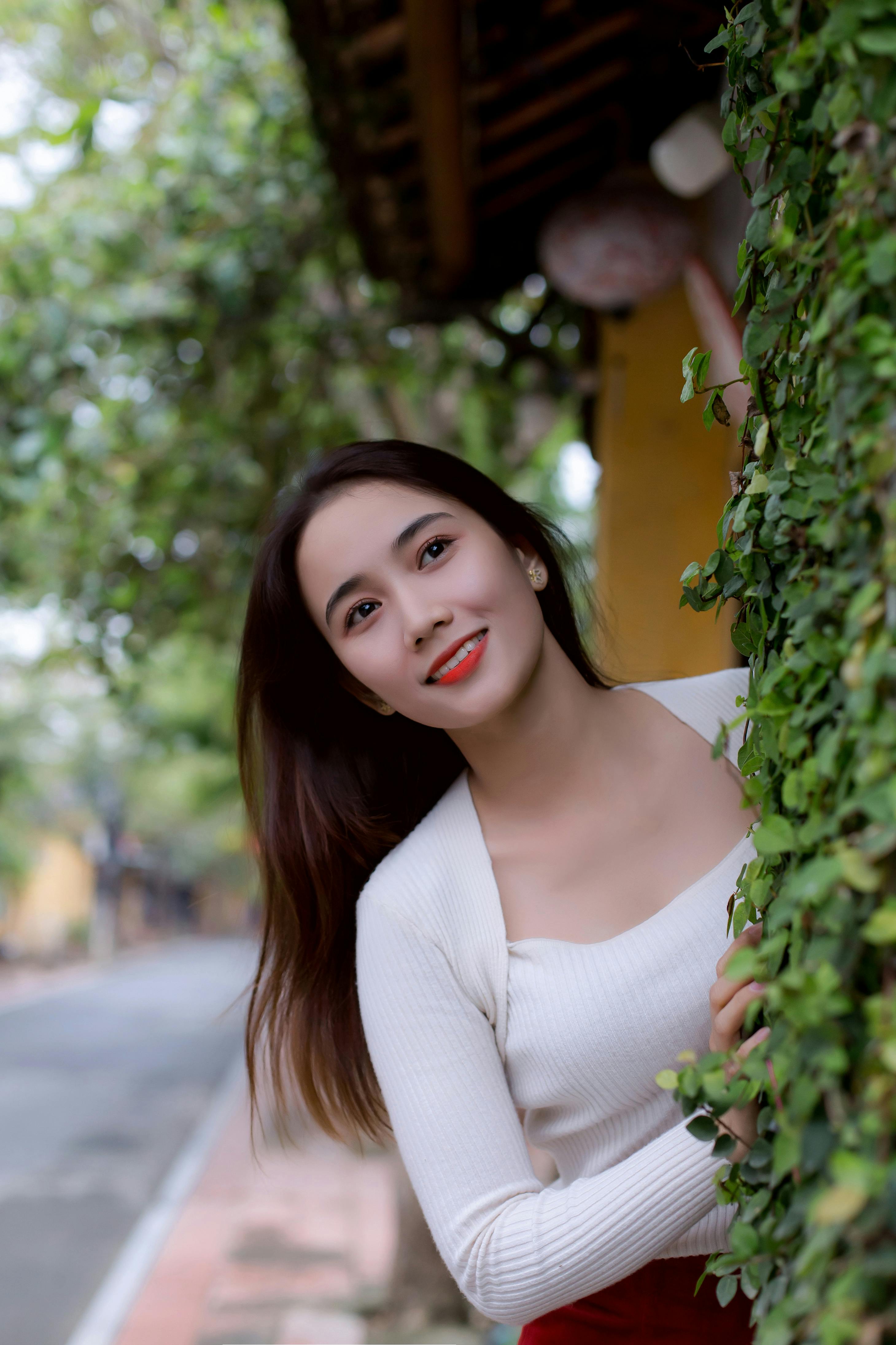 Young woman smiling, peeking from behind green foliage in Hoi An, Vietnam.