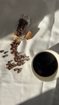 Coffee beans spill from jar beside cup of coffee on white cloth in sunlight.